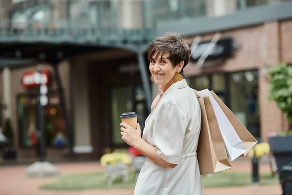 Femme âgée gaie avec les cheveux courts tenant des sacs à provisions et du café pour aller près de la sortie, à l'extérieur — Photo de stock