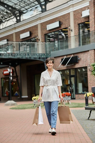 Mujer mayor complacida caminando con bolsas de compras en la calle urbana, al por menor, ancianos en la ciudad, al aire libre - foto de stock