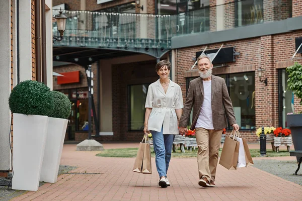 Feliz pareja de ancianos teniendo compras, hombre y mujer mayores tomados de la mano y caminando por la calle - foto de stock