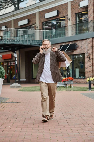 Felice e anziano uomo con la barba parlando su smartphone, tenendo borse della spesa, camminando vicino outlet — Foto stock