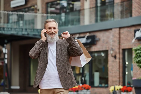 Позитивный пожилой человек с бородой разговаривает по смартфону, держит сумки с покупками, идет рядом с розеткой — стоковое фото