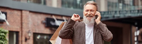 Pancarta, hombre mayor positivo con barba hablando en el teléfono inteligente, la celebración de bolsas de compras, toma de corriente - foto de stock