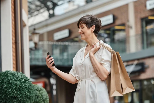 Feliz anciana usando teléfono inteligente, sosteniendo bolsas de compras y de pie cerca del centro comercial al aire libre - foto de stock