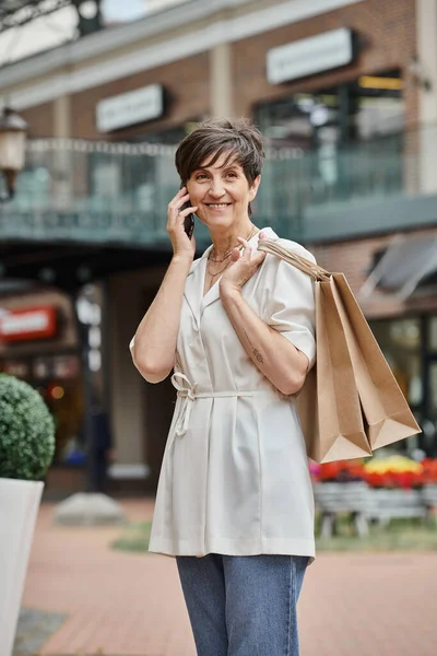 Щаслива літня жінка розмовляє по смартфону, тримає сумки для покупок і стоїть біля відкритого торгового центру — стокове фото