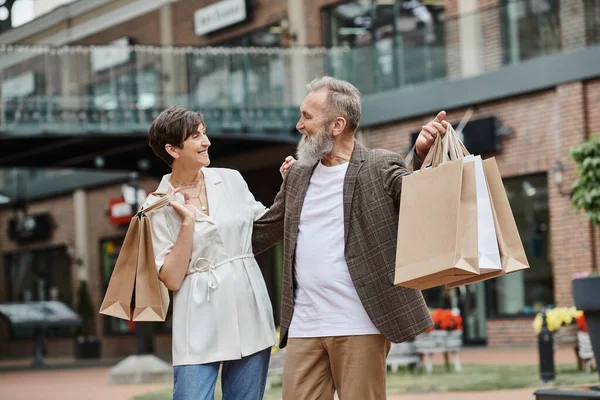 Feliz pareja sosteniendo bolsas de compras, anciano hombre y mujer mirándose, centro comercial al aire libre - foto de stock