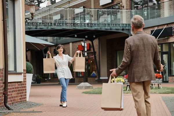 Heureuse femme âgée montrant des sacs à provisions à l'homme, mari et femme en sortie, à l'extérieur, style de vie — Photo de stock
