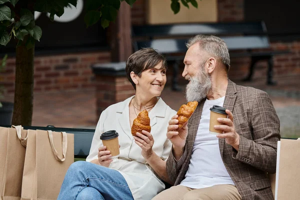 Heureux couple âgé tenant croissants et café à emporter, gobelets en papier, sacs à provisions, sortie, urbain — Photo de stock