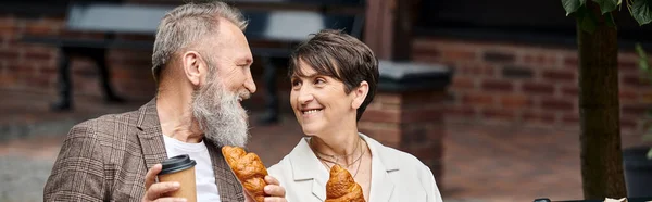 Feliz pareja de ancianos sosteniendo croissants y café para llevar, taza de papel, al aire libre, pancarta horizontal - foto de stock