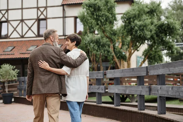 Feliz y anciano hombre y mujer abrazando y caminando juntos al aire libre, pareja mayor, romance - foto de stock