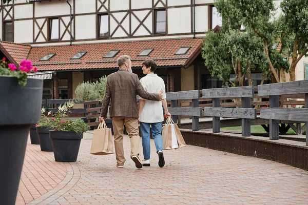 Rückansicht eines älteren Paares, das mit Einkaufstaschen spaziert, älterer Mann umarmt Frau, urbaner Lebensstil — Stockfoto