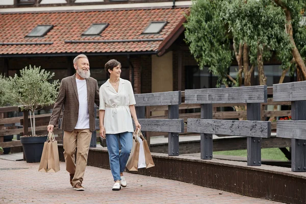 Positives Seniorenpaar geht mit Einkaufstüten, älterer Mann umarmt Frau, geht zusammen — Stockfoto