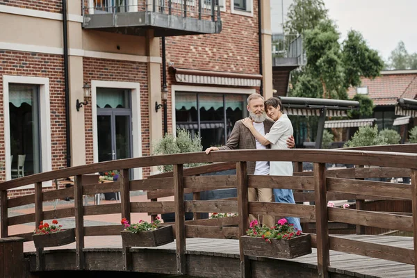 Pareja de ancianos románticos de pie juntos en el puente de madera y abrazos, el amor de edad avanzada, mira la cámara - foto de stock