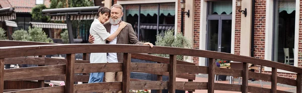 Романтическая старшая пара, стоящая вместе на деревянном мосту и обнимающая, пожилая любовь, плакат — стоковое фото