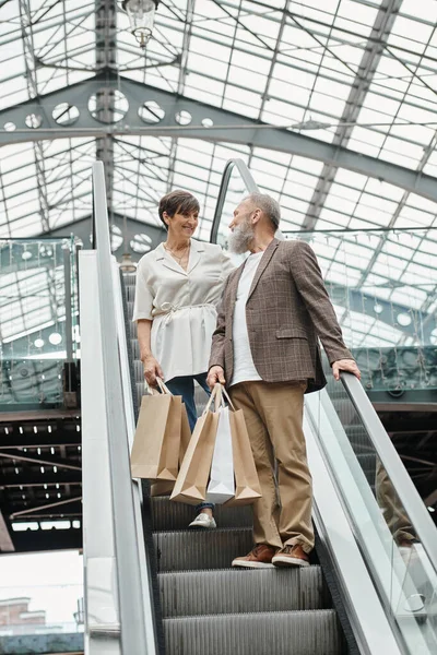 Felice anziano uomo e donna in piedi sulla scala mobile, borse della spesa, guardarsi l'un l'altro nel centro commerciale — Foto stock