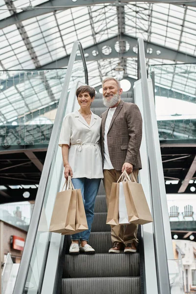 Allegro anziano uomo e donna in piedi sulla scala mobile, borse della spesa, guardando la fotocamera nel centro commerciale — Foto stock