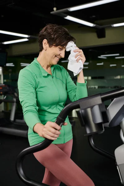 Sénior actif, femme âgée heureuse en vêtements de sport essuyant la sueur avec une serviette, machine d'exercice stepper — Photo de stock
