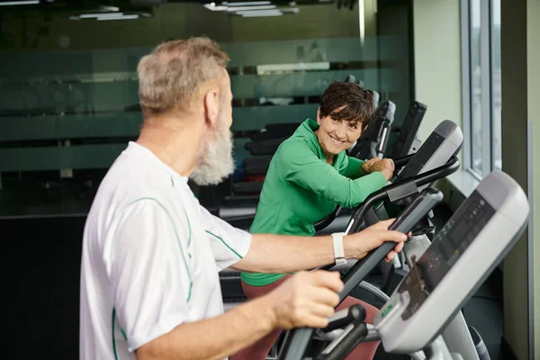 Cheerful woman looking at elderly man, husband and wife exercising in gym, active seniors, sport — Stock Photo