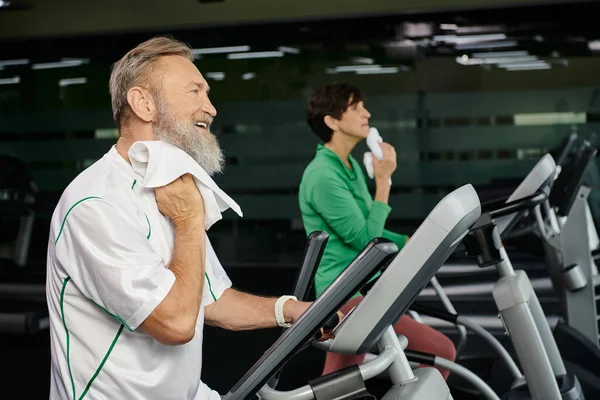 Senior and bearded man wiping sweat with towel, blurred woman on background, exercising in gym — Stock Photo