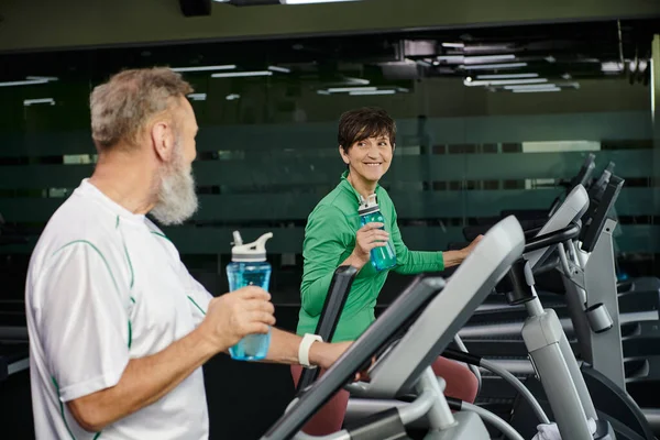 Cheerful woman looking at elderly man, husband and wife working out in gym, holding sports bottles — Stock Photo