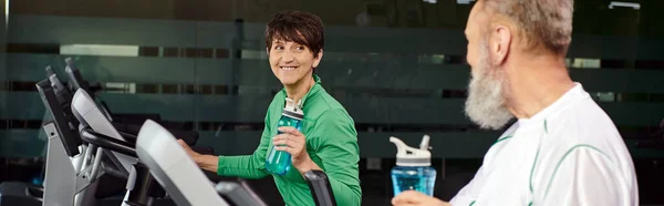 Glückliche Frau blickt älteren Mann, Mann und Frau im Fitnessstudio an, hält Sportflaschen, Banner — Stockfoto