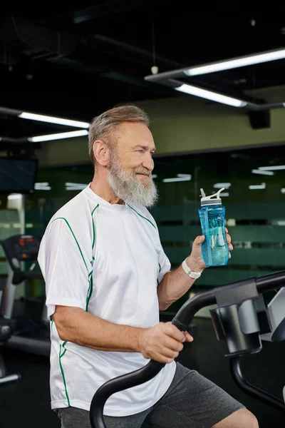 Счастливый пожилой и бородатый мужчина, держа в руках спортивную бутылку с водой и отводя взгляд, спорт, тренажерный зал, тренировка — стоковое фото
