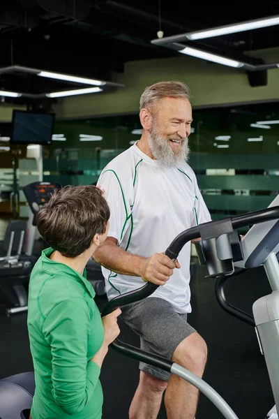 Hombre mayor feliz y barbudo ejercicio cerca de la mujer en el gimnasio, marido y mujer, personas mayores activas, deporte — Stock Photo