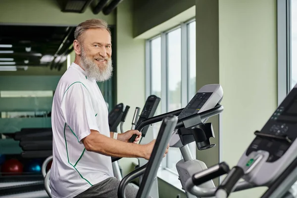 Happy bearded man working out on exercise machine, elderly in gym, active senior, fitness and sport — Stock Photo