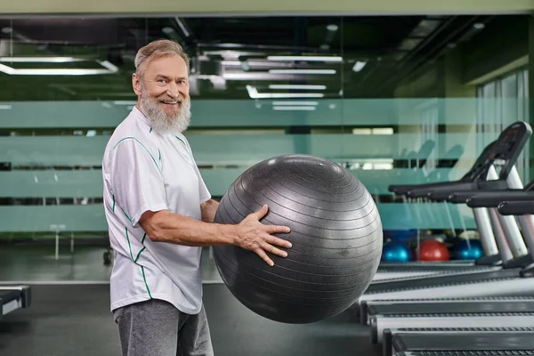 Glücklicher älterer Herr mit Fitnessball im Fitnessstudio, Workout, Fitness und Sport, schaut in die Kamera, aktiv — Stockfoto