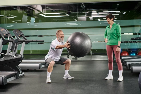 Excité homme âgé faisant squats avec balle de remise en forme près de la femme, mari et femme dans la salle de gym, couple, sport — Photo de stock