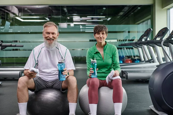 Sporty elderly couple, happy man and woman sitting on fitness balls, holding bottles with water — Stock Photo