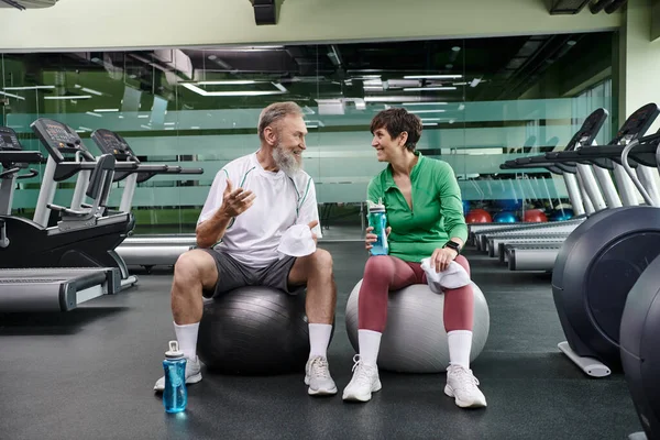 Sporty elderly couple, cheerful man and woman sitting on fitness balls, holding bottles with water — Stock Photo