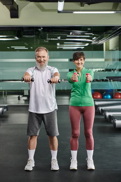 Pareja de ancianos positivos, feliz hombre y mujer entrenando con pesas, marido y mujer en el gimnasio - foto de stock