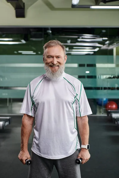 Feliz y barbudo anciano sonriendo y mirando a la cámara, haciendo ejercicio con mancuernas en el gimnasio - foto de stock