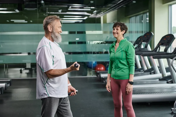 Mujer feliz mirando al marido barbudo haciendo ejercicio con pesas en el gimnasio, personas mayores activas, estilo de vida — Stock Photo