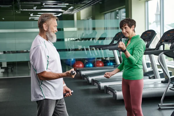 Femme heureuse regardant homme barbu travaillant avec haltères dans la salle de gym, personnes âgées actives, mode de vie — Photo de stock