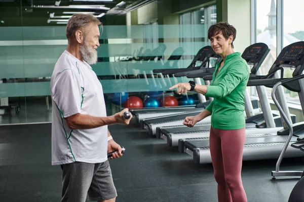 Femme heureuse pointant vers l'homme barbu travaillant avec des haltères dans la salle de gym, personnes âgées actives, mode de vie — Photo de stock