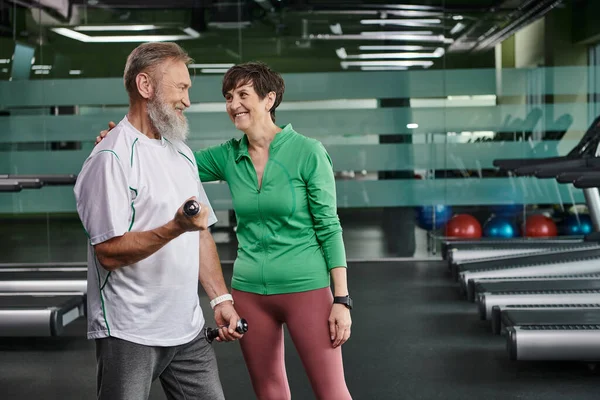 Femme heureuse encourageant homme barbu travaillant avec haltères dans la salle de gym, personnes âgées actives, mode de vie — Photo de stock
