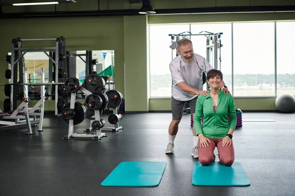Homme âgé heureux avec barbe encourageant femme dans la salle de gym, actif, mari et femme, tapis de fitness — Photo de stock