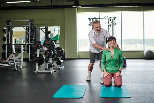 Happy elderly man with beard encouraging woman in gym, active, husband and wife, fitness mats — Stock Photo