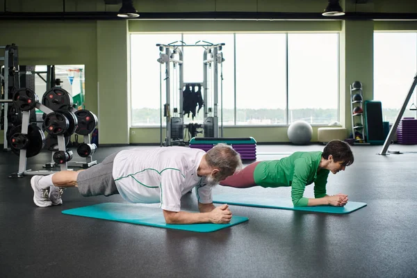 Elderly man and woman doing plank on fitness mats, active seniors exercising on in gym, healthy life — Stock Photo