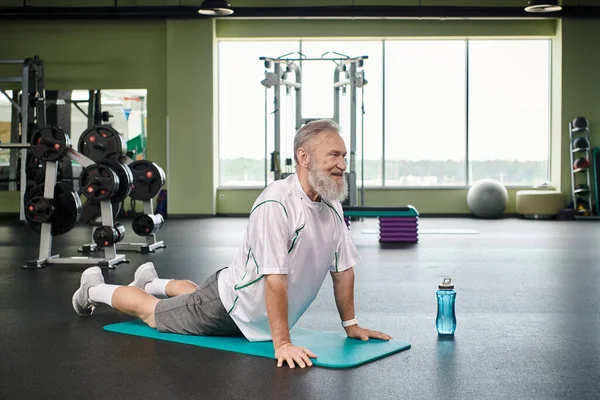Homme âgé heureux avec barbe étirant en arrière sur tapis de fitness, senior actif, vibrant et en bonne santé — Photo de stock