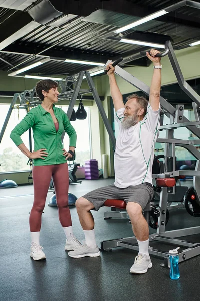 Strong and elderly man with beard working out on exercise machine near happy woman, gym, active — Stock Photo
