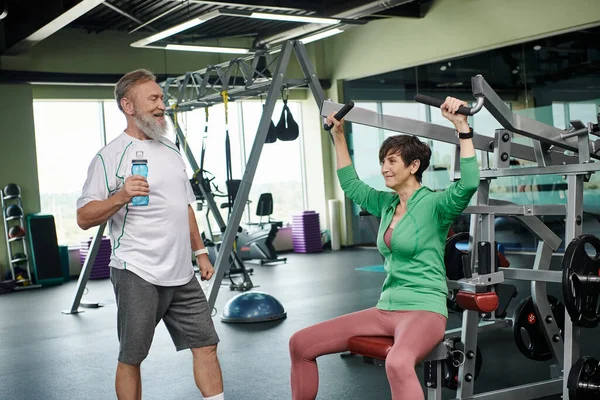 Mujer atlética y anciana haciendo ejercicio en la máquina de ejercicio cerca de hombre feliz, gimnasio, botella con agua — Stock Photo