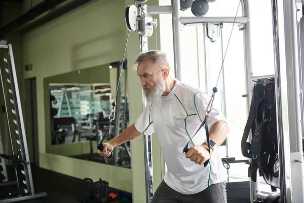 Anciano motivado con barba haciendo ejercicio en la máquina de ejercicio en el gimnasio, atleta, senior activo - foto de stock