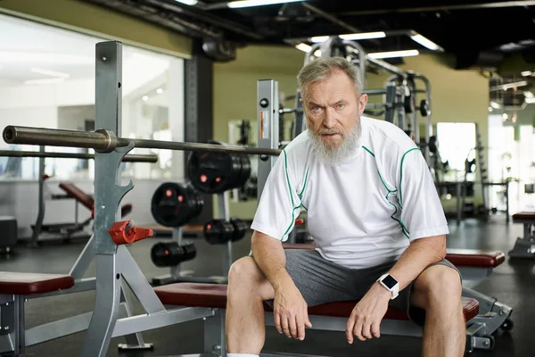 Anciano cansado con barba mirando a la cámara después del entrenamiento, máquina de ejercicios en el gimnasio, atleta - foto de stock