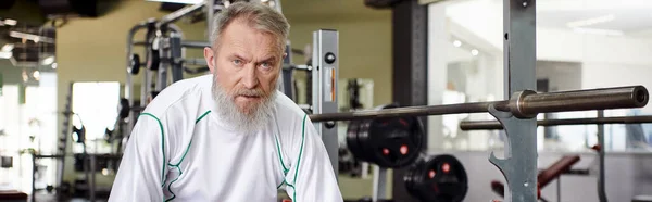 Tired elderly man with beard looking at camera after workout, exercise machine in gym, banner — Stock Photo