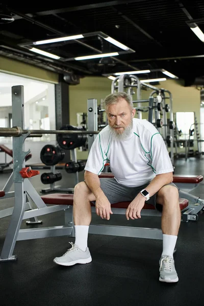 Müder älterer Mann mit Bart blickt nach dem Training in die Kamera, sitzt im Fitnessstudio auf einem Trainingsgerät — Stockfoto