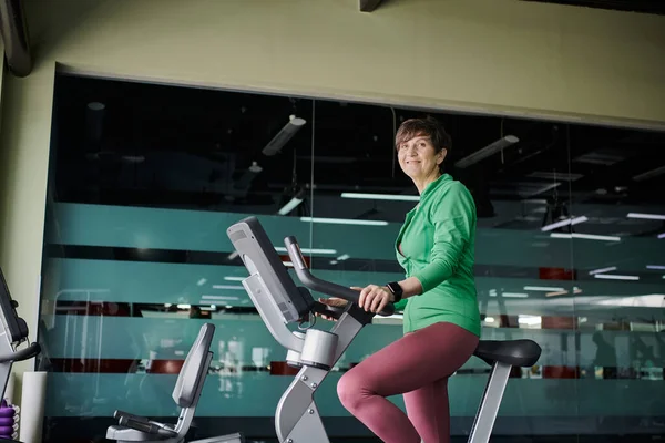 Femme âgée heureuse avec les cheveux courts travaillant sur vélo d'exercice dans la salle de gym, active, motivation — Stock Photo