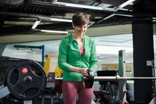Femme âgée souriante et debout près de l'haltère avec plaque de poids, séance d'entraînement en salle de gym, motivation — Stock Photo