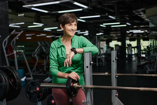 Heureuse femme âgée souriante et debout près de l'haltère, plaques de poids, séance d'entraînement en salle de gym, motivation — Photo de stock